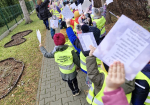 Dzieci ustawione w parach przed przedszkolem w kamizelkach odblaskowych podnoszą do góry przygotowane przez siebie kartki świąteczne.