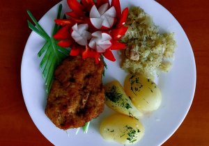 Na stoliku leży talerz, na którym znajdują się: ziemniaki, kotlet schabowy, gotowana kapusta, rzodkiewka i papryka.