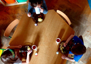Dzieci siedzą przy stoliku i jedzą drugie danie. Dziewczynka pije kompot.