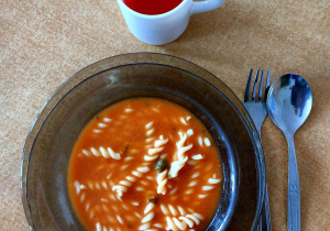 Na stoliku leży talerz z zupą pomidorową oraz sztućce. Obok stoi kubek z kompotem.
